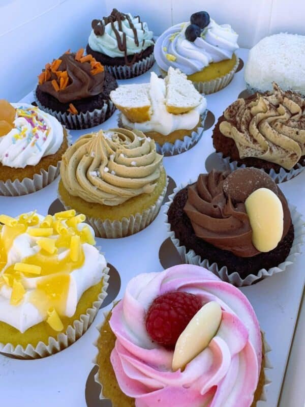 A selection of assorted cupcakes with colourful toppings including chocolate, raspberry, lemon, and caramel, displayed in a white box.