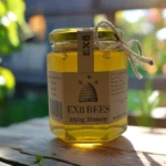 A close-up photo of a 250g jar of EX8 BEES honey on a beehive, with bees crawling on the honeycomb and label. The jar has a kraft paper label and rustic twine, and the honey inside glows golden in the sunlight.